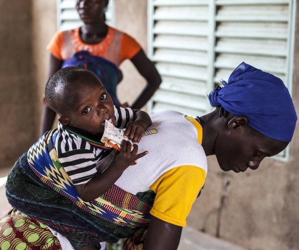 A young boy, on his mother's back, eats Plumpy'Nut to recover from malnutrition.