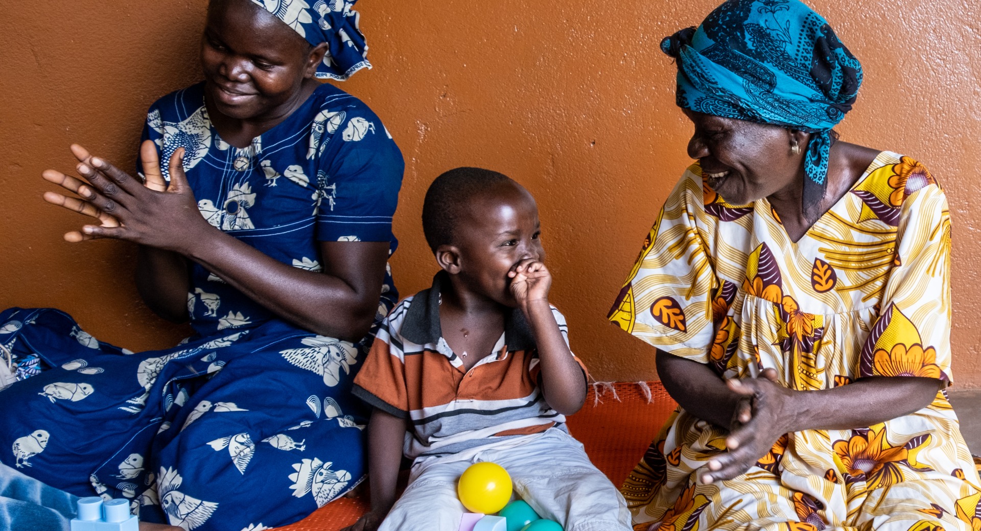 Un enfant handicapé par une malnutrition sévère à un jeune âge reçoit une formation en motricité au Centre de Réhabilitation pour les Handicapés Physiques (CRHAM) soutenu par ACF. Ici, il est entouré de la mère d'un autre enfant comme lui et de sa grand-mère. Bangui, RCA. 2022