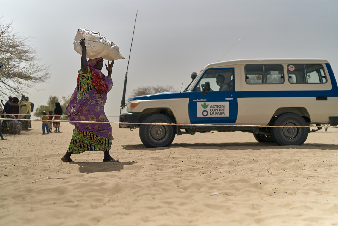 A woman walks through dry, sandy land with a large bag above her head. An Action Against Hunger truck is in the background.