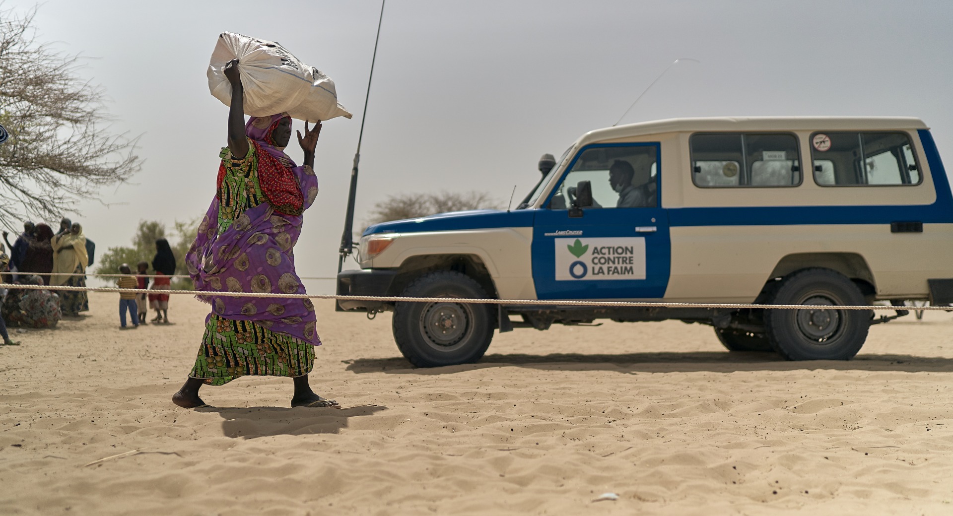 A woman walks through dry, sandy land with a large bag above her head. An Action Against Hunger truck is in the background.