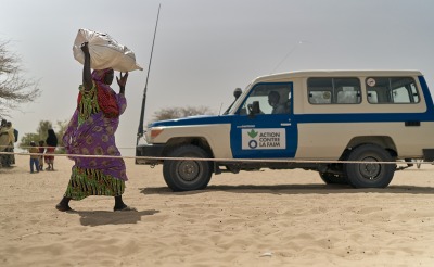 A woman walks through dry, sandy land with a large bag above her head. An Action Against Hunger truck is in the background.