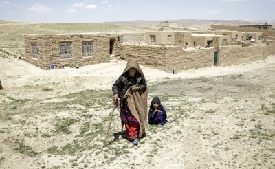 A mother walks away from her home with the help of a cane. Her daughter crouches behind her.