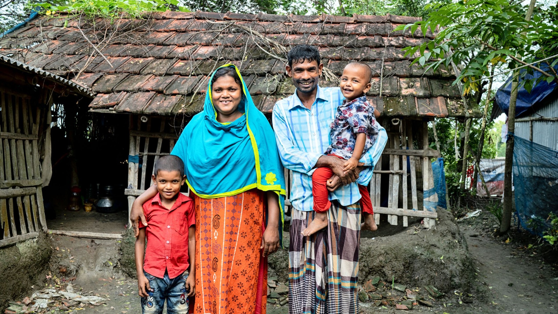 A mother and a father stand smiling with their two sons outside of their home in Bangladesh.