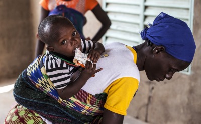 A young boy, on his mother's back, eats Plumpy'Nut to recover from malnutrition.