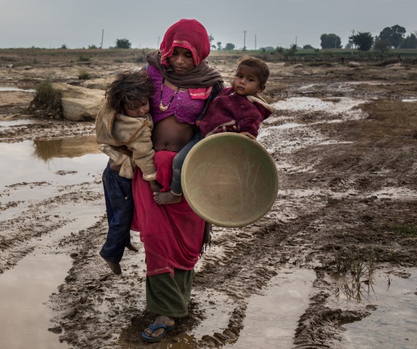 A mother carries her two children over a muddy road.